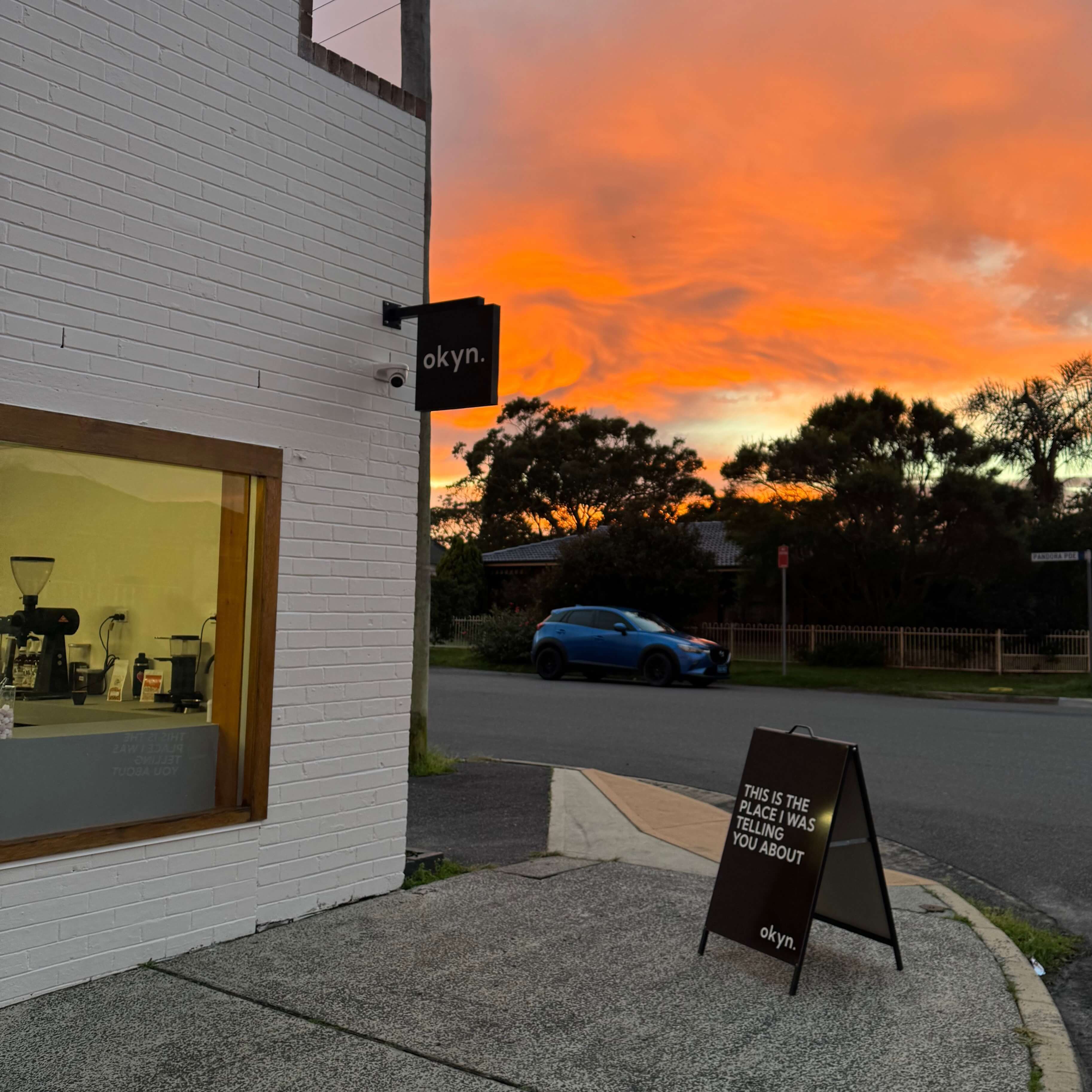 A cafe on the corner with a vibrant orange sunrise rolling in the sky. On the pavement sits a A-framed sign that says "This is the place I was telling you about"